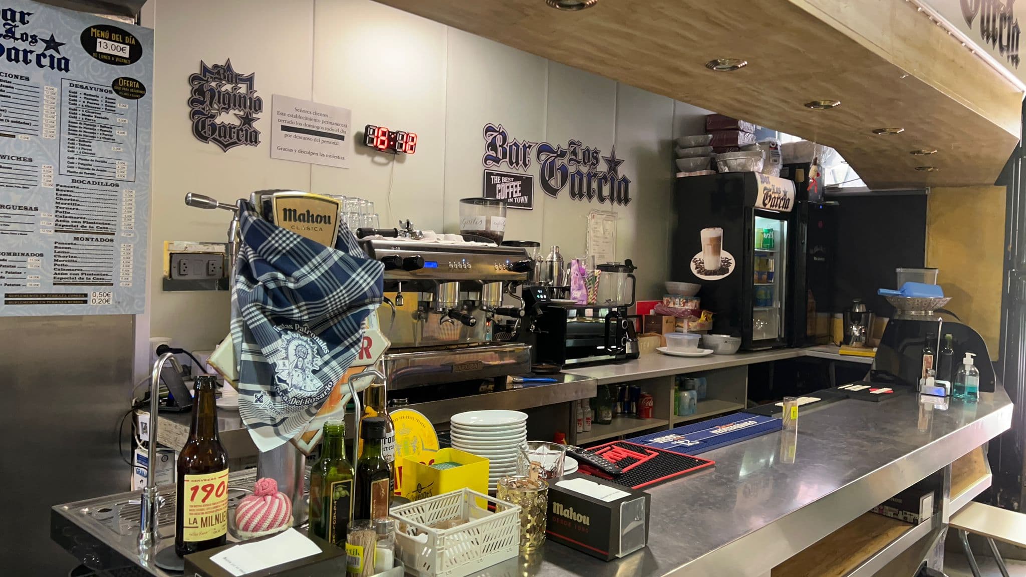 Interior de bar con barra metálica, cafetera, grifos de cerveza y carteles de menús.