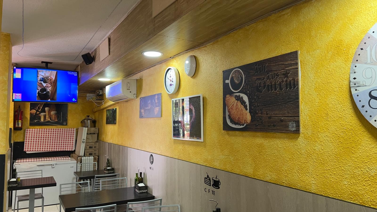 Cafetería con paredes amarillas, cuadros decorativos, mesas negras, sillas metálicas y un televisor encendido.