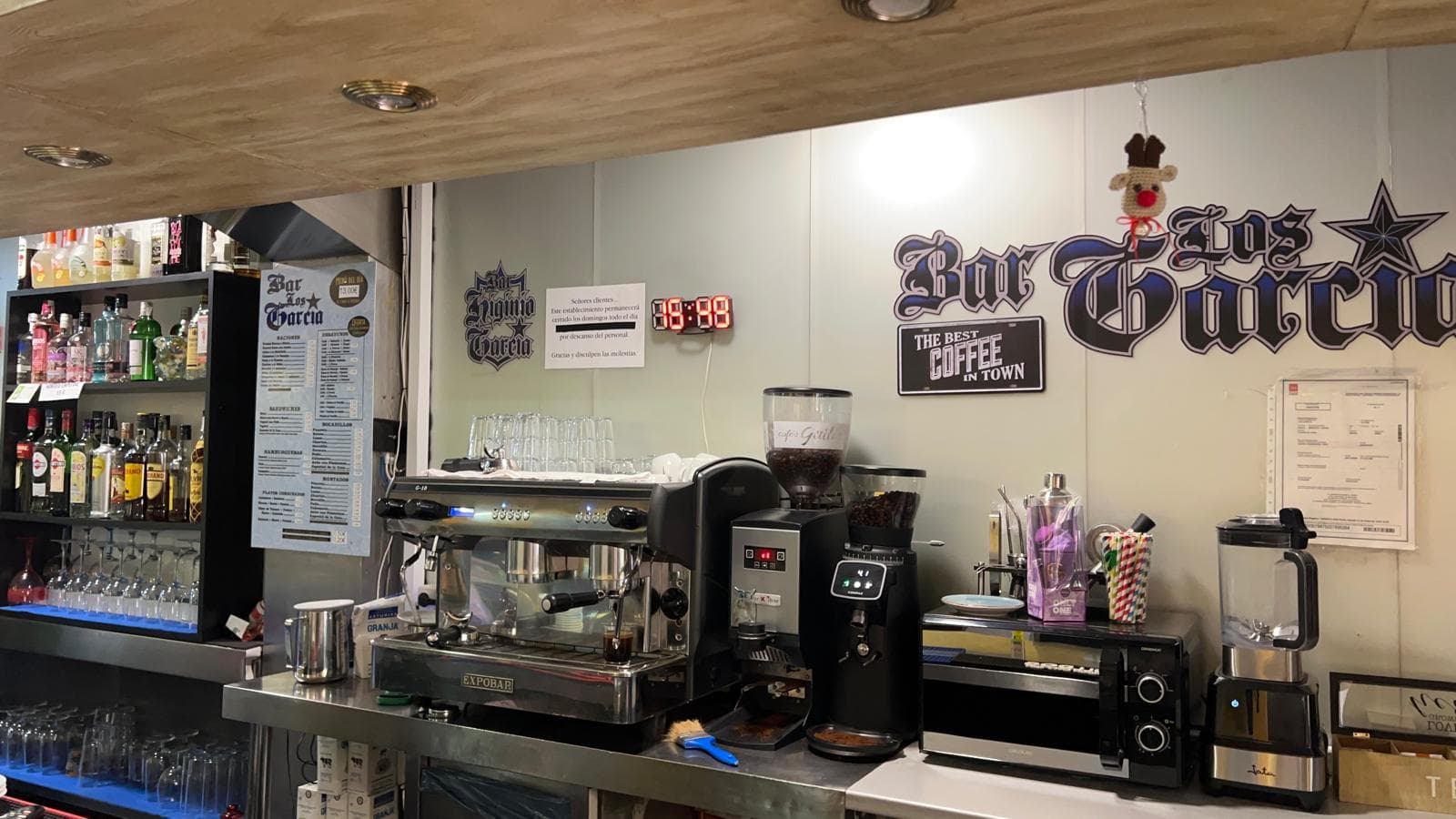 Barra de bar con máquina de café, molinillos y estanterías con botellas de licores.